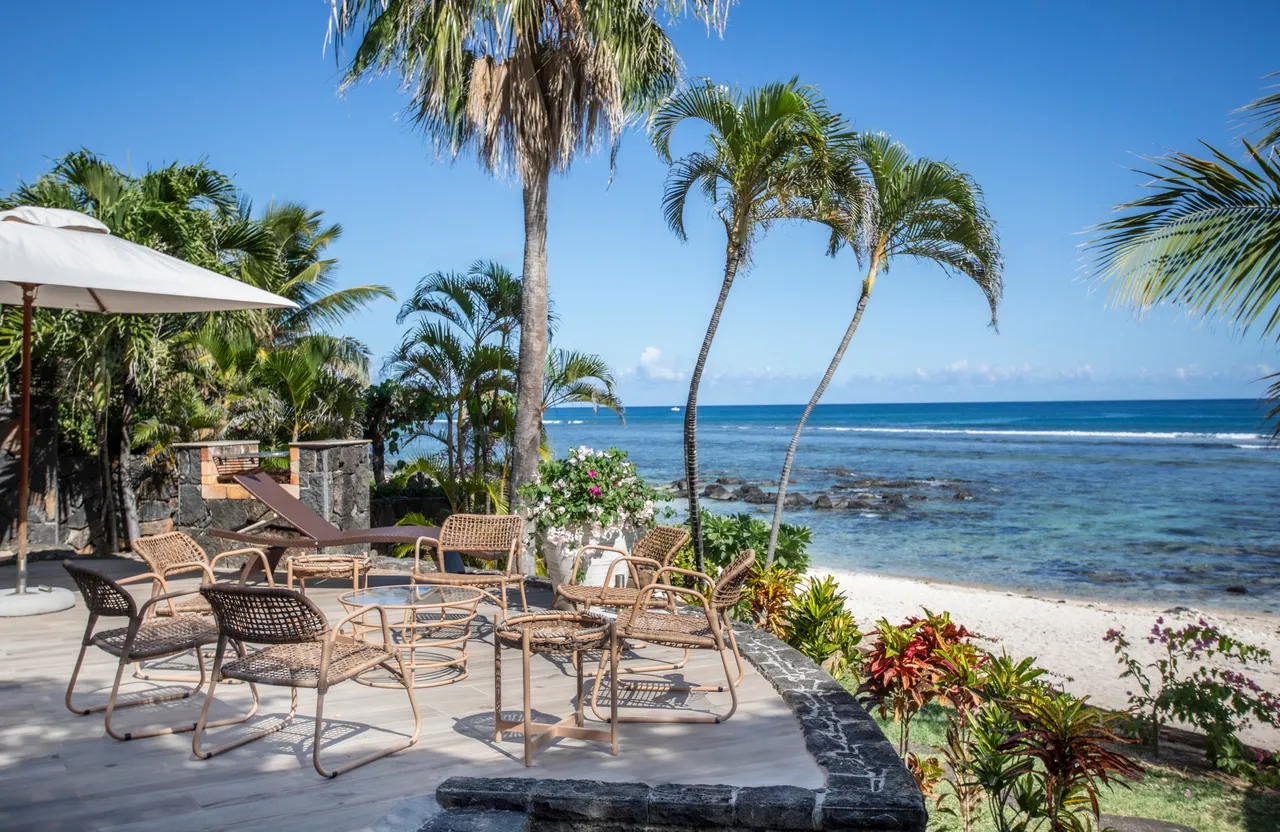 Terrasse avec mobilier en rotin et parasol, vue directe sur la mer. Végétation tropicale encadre l'espace extérieur.