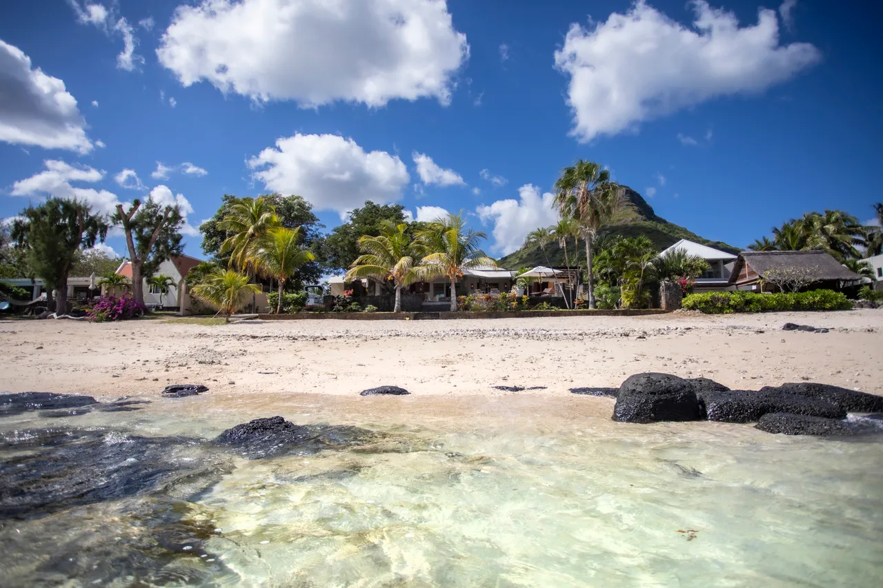 Plage de sable avec eau turquoise, vue dégagée sur les montagnes. Végétation tropicale en arrière-plan.