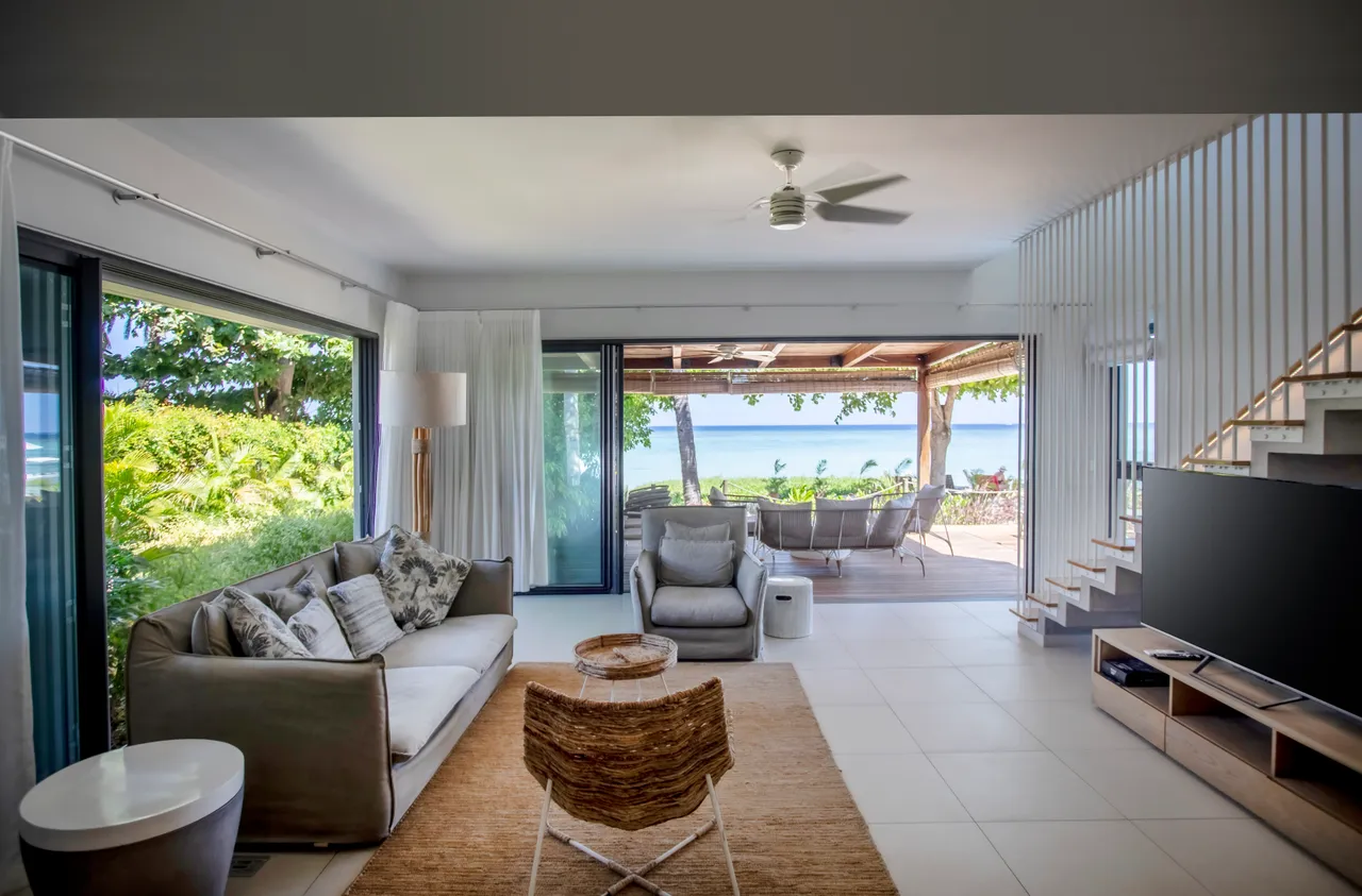 Salon avec de grandes baies vitrées donnant sur l'extérieur. Mobilier en teintes claires et décor minimaliste. Vue sur la terrasse et la piscine en arrière-plan. Éclairage naturel grâce aux ouvertures sur les deux côtés.