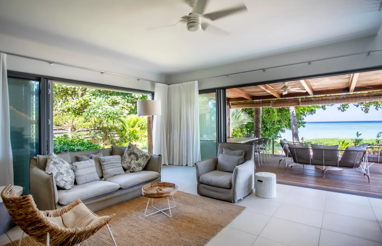 Sol en carrelage clair avec un agencement minimaliste et mobilier en teintes douces. Canapé confortable et fauteuils accueillants, offrant une vue sur la terrasse. Grandes baies vitrées qui laissent entrer la lumière naturelle et ouvrent sur l'extérieur. Décoration sobre avec des touches de textile en ton neutre. Terrasse en bois en continuité avec le salon.