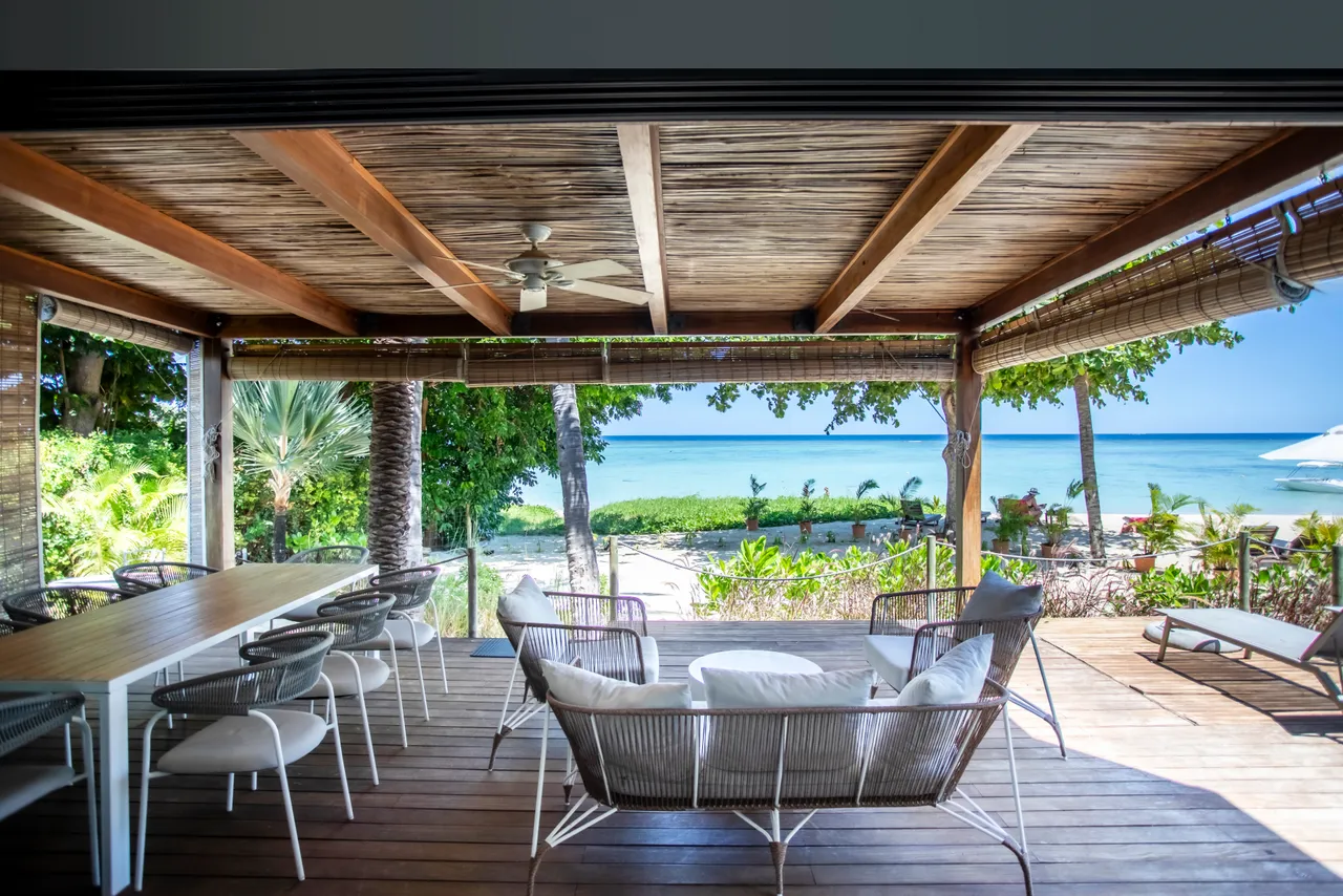 Terrasse en bois avec mobilier en rotin et vue sur la mer.  
Éclairage naturel et ventilation grâce aux ouvertures latérales.  
Table à manger spacieuse, idéale pour des repas en extérieur.  
Chaises confortables disposées autour d'un espace convivial sur la terrasse.  
Plantes tropicales ajoutent une touche de verdure au cadre extérieur.  
Bord de mer