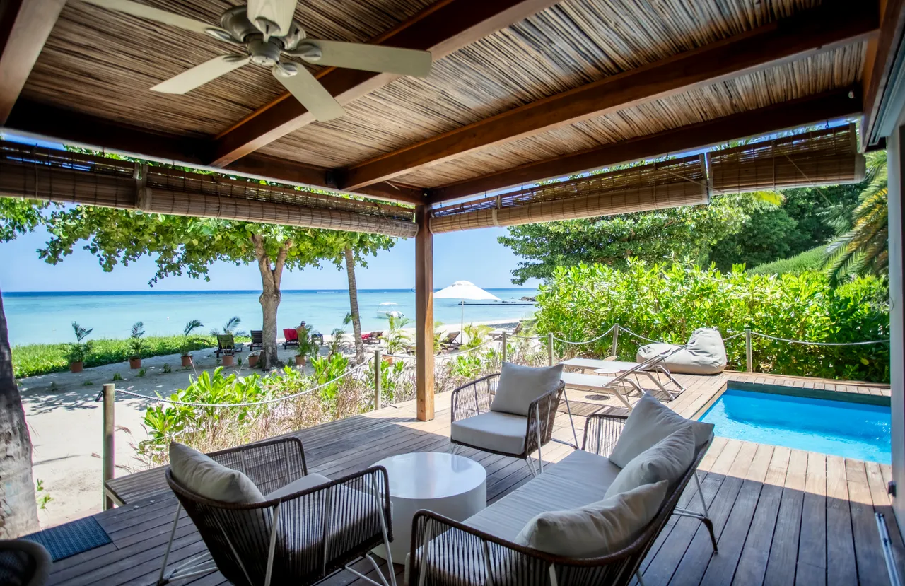 Bois naturel et textures douces sur la terrasse avec vue dégagée. Espace de détente avec fauteuils confortables et table basse. Piscine privée visible à proximité, entourée de verdure. Atmosphère tranquille offrant un accès direct à la plage. Ventilation assurée par le plafond ventilé.