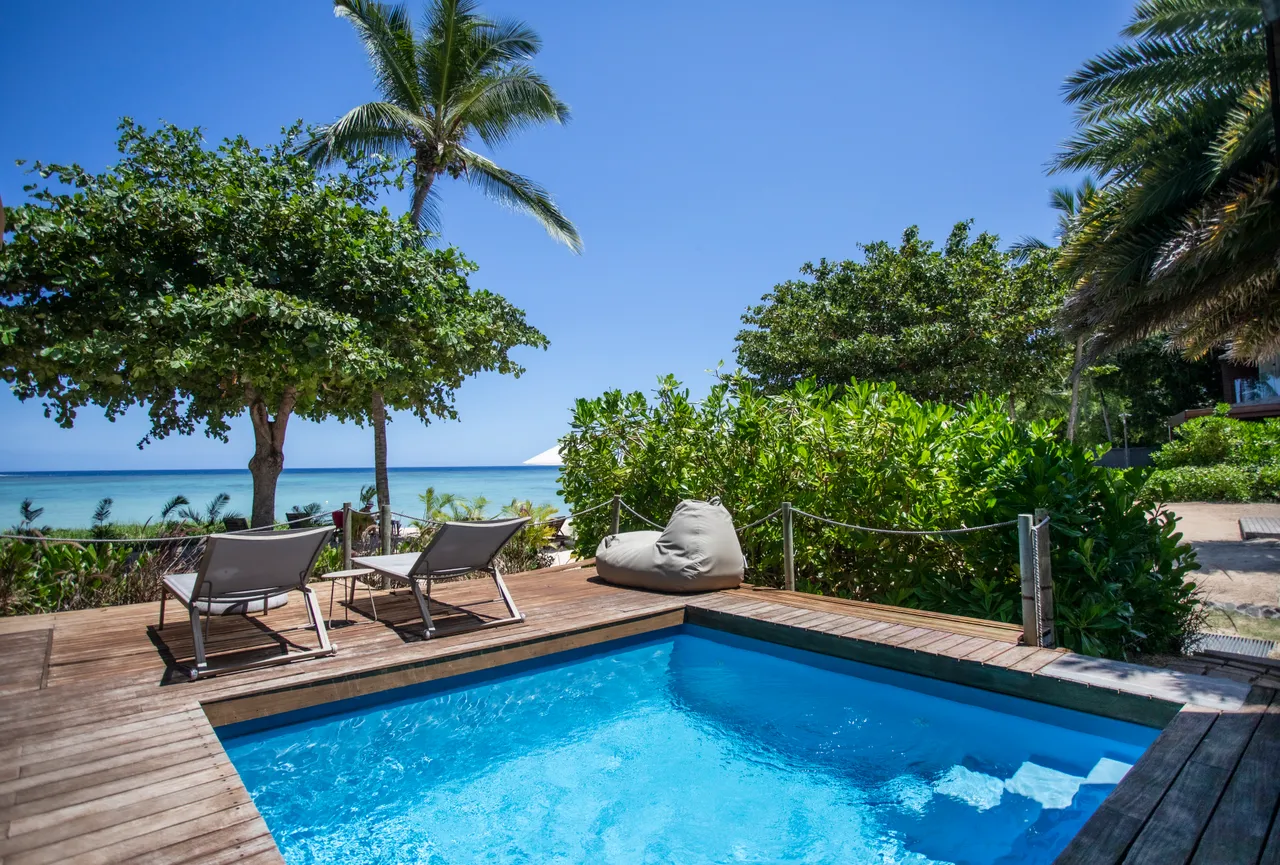 Teintes bleues et textures naturelles autour de la piscine privée. Chaises longues sur la terrasse en bois, face à la mer. Végétation luxuriante encadre l'espace extérieur. Accès direct à la plage depuis la zone de détente.