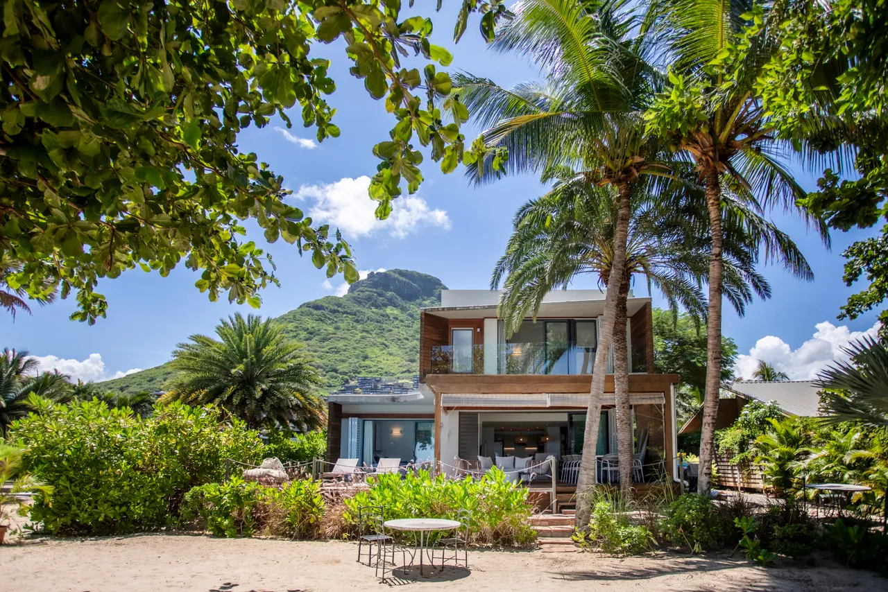 Villa avec architecture moderne entourée de verdure tropicale. Accès direct à la plage depuis le jardin. Façade en bois et grandes baies vitrées offrant une vue sur l'environnement extérieur. Palmier en arrière-plan ajoutant une touche naturelle à l'espace.