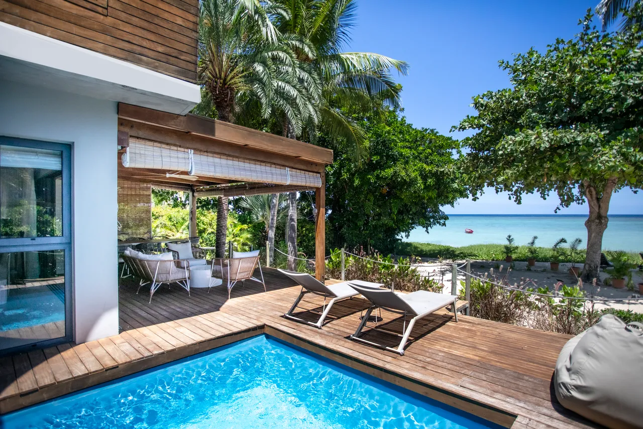 Espace de détente avec chaises longues autour de la piscine privée. Accès direct à la plage depuis la terrasse. Ombre naturelle fournie par les palmiers environnants. Coin repas avec mobilier en rotin, offrant une vue sur la mer. Terrasse en bois intégrée dans un environnement verdoyant.