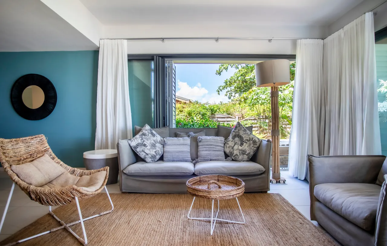 Salon avec canapé gris et fauteuil en osier. Grandes baies vitrées apportent une vue sur l'extérieur. Tapis en jute ajoutant une texture naturelle au sol. Décoration simple avec coussins et lampe sur pied. Espace lumineux grâce aux ouvertures.