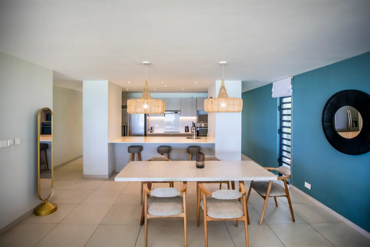 Teintes neutres et textures claires dans l'espace repas. Table spacieuse entourée de chaises confortables. Éclairage suspendu en rotin ajoutant une touche naturelle. Cuisine ouverte offrant un accès direct à la salle à manger. Miroir décoratif apportant de la profondeur à la pièce.
