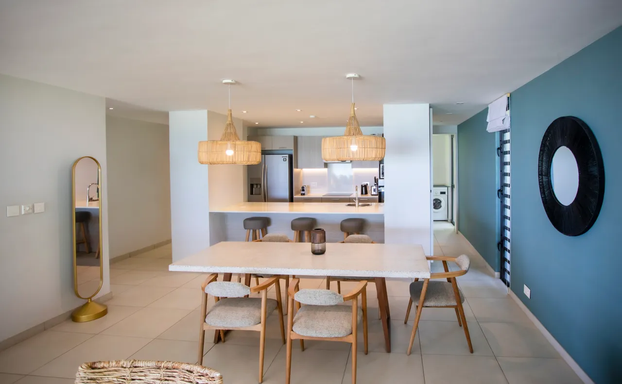 Espace repas lumineux avec table en marbre et chaises en bois. Éclairage suspendu en rotin ajoutant une touche naturelle. Cuisine ouverte adjacente pour un accès facilité. Mur en teintes claires créant une ambiance aérée. Miroir décoratif offrant une perception de profondeur.