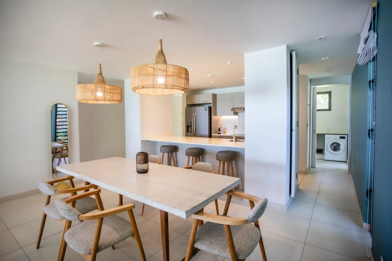 Espace repas lumineux avec table en marbre et chaises en bois clair. Luminaires en rotin ajoutent une ambiance chaleureuse. Cuisine ouverte à proximité pour une circulation aisée. Miroir décoratif agrandissant visuellement la pièce. Sol en carrelage clair offrant une surface fonctionnelle.