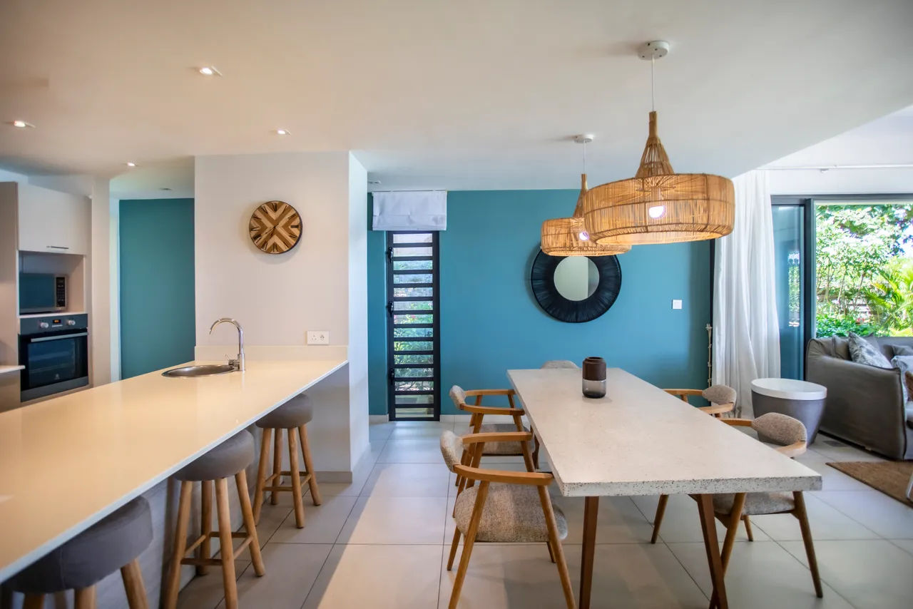 Espace repas moderne avec table en pierre et chaises en bois. Bar à petit-déjeuner adjacent avec tabourets confortables. Murs en teintes claires ajoutent une ambiance lumineuse. Accès immédiat à la terrasse verdoyante depuis la salle à manger. Éléments décoratifs en rotin apportent une touche naturelle.