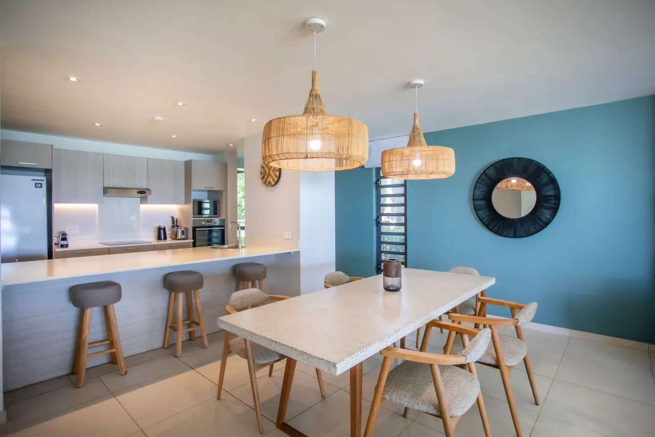 Espace repas avec table en pierre et chaises en bois clair. Éclairage suspendu en rotin ajoutant une ambiance chaleureuse. Cuisine ouverte adjacente pour une circulation aisée. Murs en teintes claires créant une atmosphère lumineuse. Miroir décoratif apportant de la profondeur à la pièce.