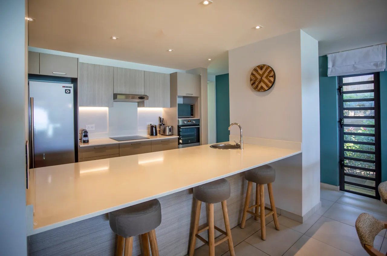 Teintes claires et surfaces lisses dans une cuisine moderne. Bar à petit-déjeuner avec tabourets en bois. Équipements intégrés offrant un espace fonctionnel. Fenêtres laissant entrer la lumière naturelle. Rangements pratiques pour une cuisine bien agencée.