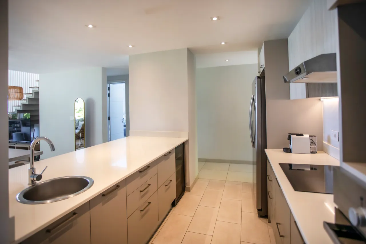Cuisine moderne avec des surfaces claires et un bar à petit-déjeuner. Électroménagers intégrés pour un usage pratique. Éclairage doux apportant une ambiance agréable. Rangements fonctionnels optimisant l'espace disponible. Fenêtres laissant entrer la lumière naturelle.