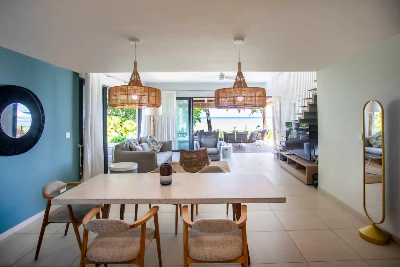 Espace repas avec table en pierre et chaises en bois. Luminaires en rotin apportent une ambiance naturelle. Accès direct à la terrasse depuis la salle à manger. Éléments décoratifs en rotin ajoutent une touche contemporaine. Grandes ouvertures offrant une vue sur l'extérieur.