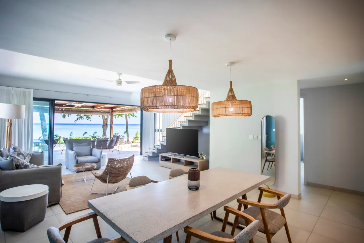 Espace repas avec table en pierre et chaises en bois clair. Luminaires en rotin créent une ambiance naturelle. Grandes baies vitrées offrant une vue sur le jardin. Accès direct à la terrasse ombragée. Décoration simple et contemporaine avec un miroir en mur.