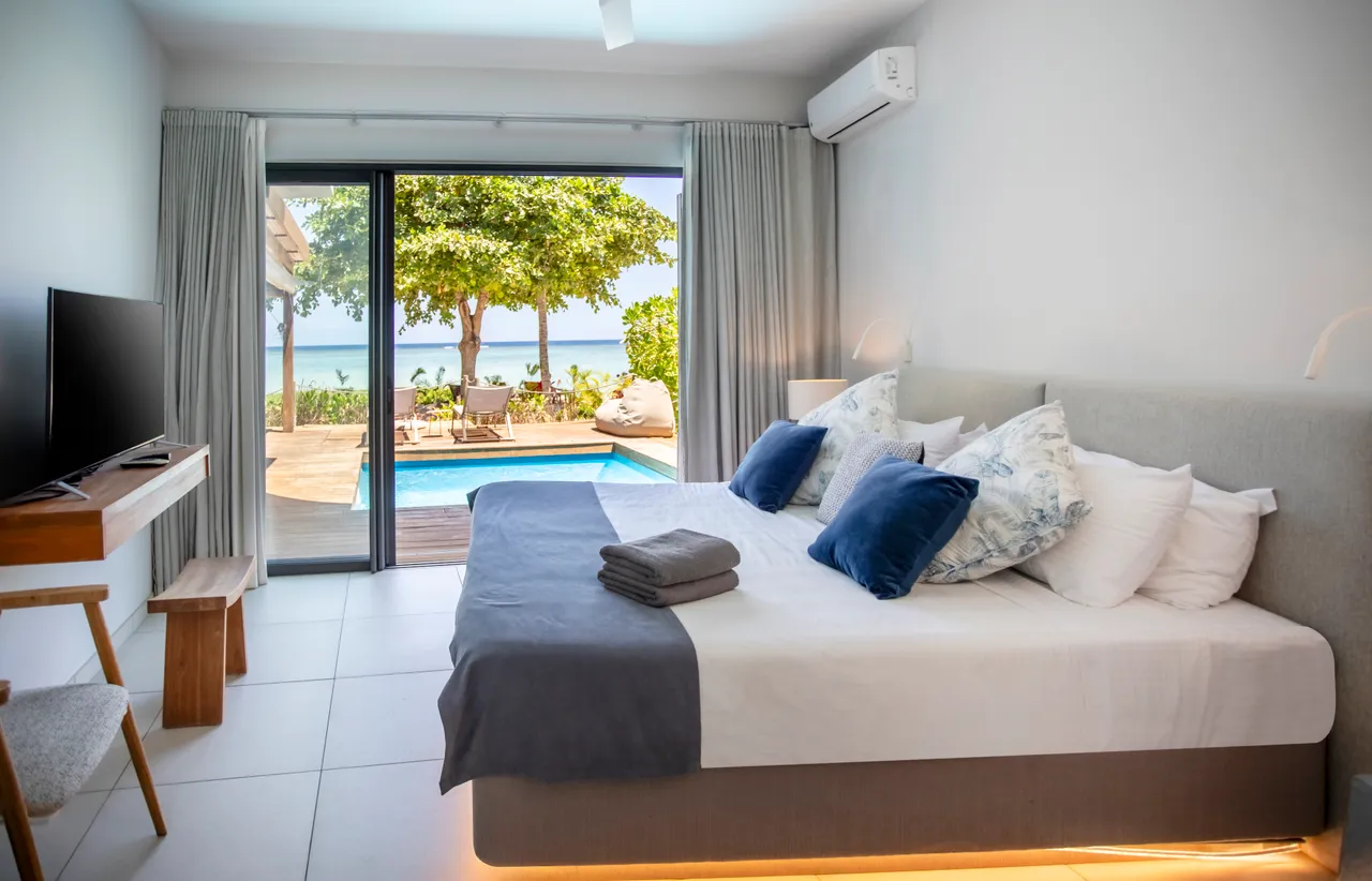 Chambre avec grandes fenêtres offrant une vue sur la piscine et la mer. Lit double confortable et décoration épurée. Accès direct à l'extérieur depuis la pièce.