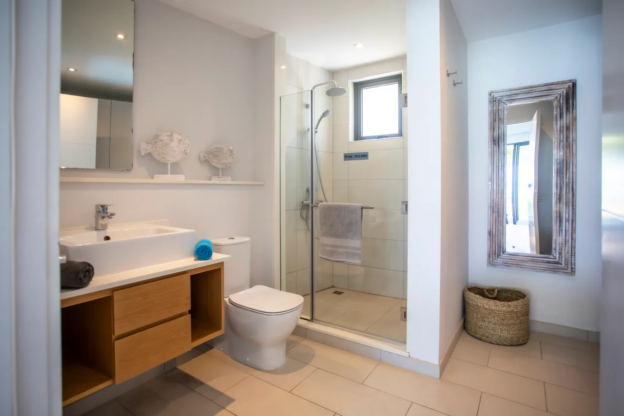 Salle de bain avec douche à l'italienne et mobilier en bois. Éclairage doux et miroir décoratif ajoutant de la profondeur. Accessoires pratiques pour un usage quotidien. Ambiance épurée avec carrelage clair.