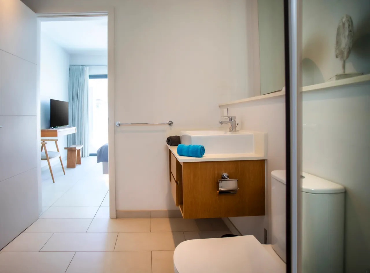 Salle de bain avec lavabo sur meuble en bois et douche à l'italienne. Éclairage naturel provenant d'une fenêtre. Rangements pratiques intégrés pour un usage quotidien. Carrelage clair apportant une ambiance épurée.