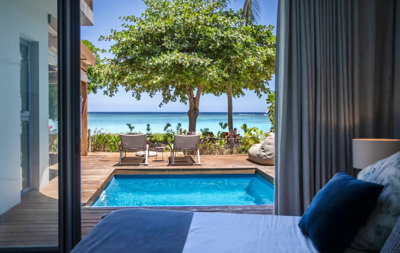 Chambre avec accès direct à la piscine et vue sur la mer. Éclairage naturel grâce aux grandes fenêtres. Mobilier sobre et décor contemporain avec touches de couleur. Terrasse en bois offrant une atmosphère extérieure agréable.