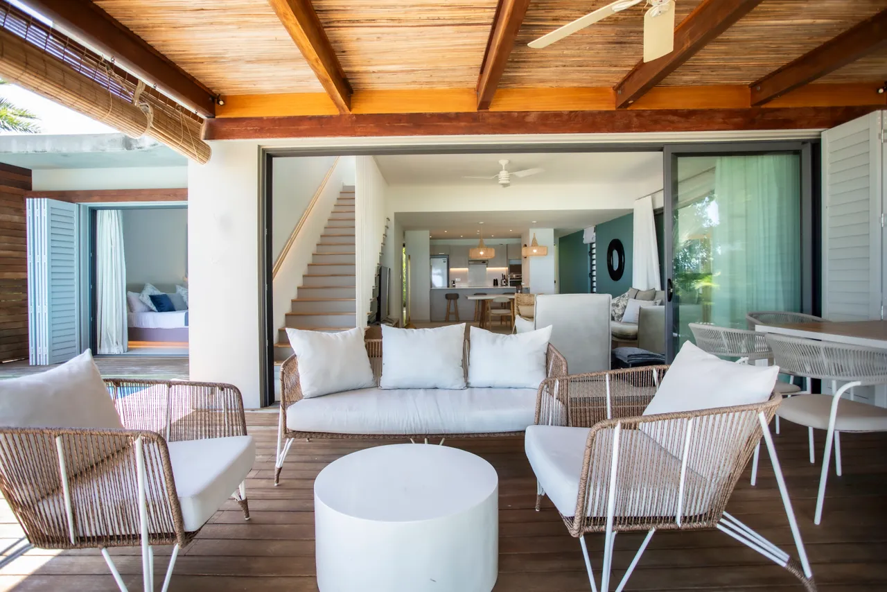 Terrasse en bois avec mobilier d'extérieur en rotin. Espace convivial pour se détendre. Accès direct à l'intérieur de la villa. Grandes baies vitrées offrant une vue sur la piscine. Éclairage naturel grâce à l'orientation de la terrasse.