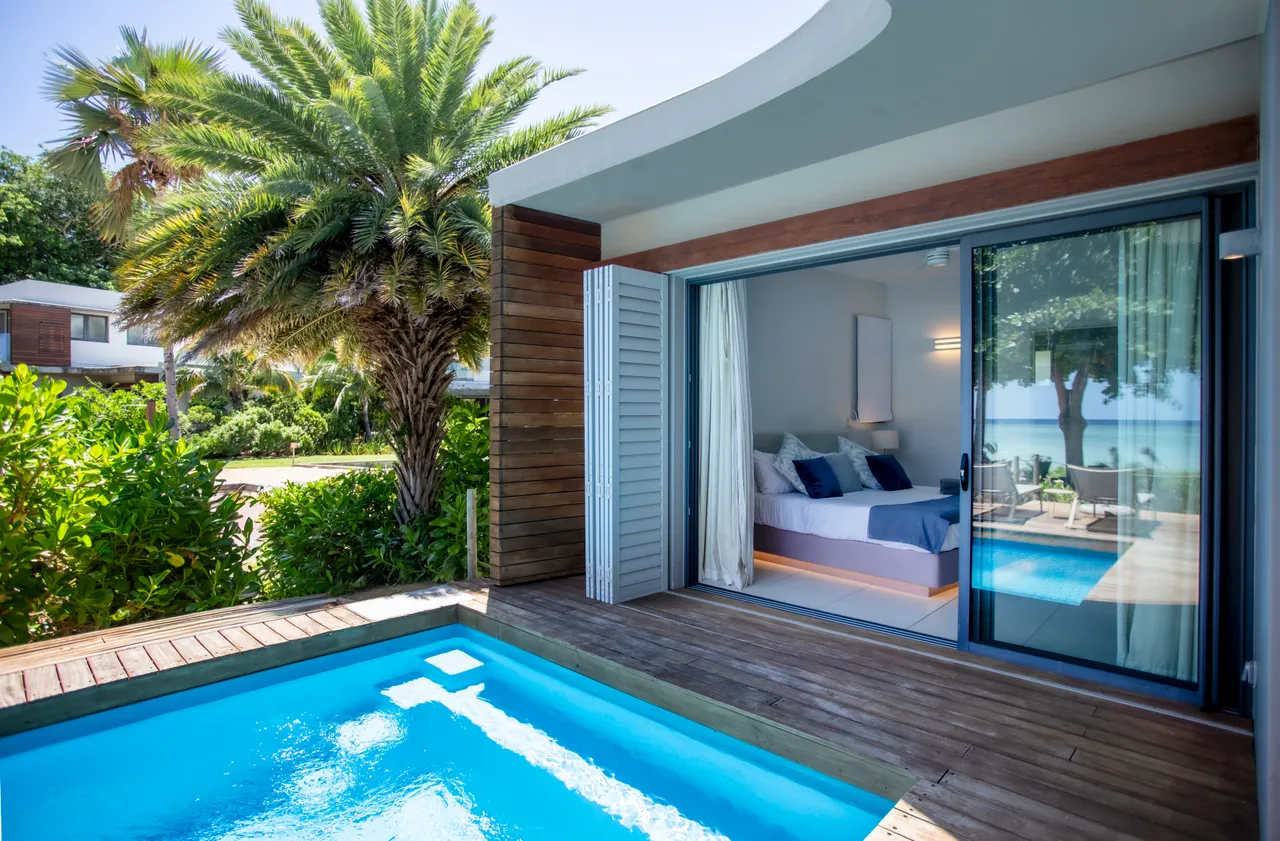 Teintes azurées de la piscine bordée de bois. Accès direct à une chambre lumineuse. Grandes baies vitrées offrant une vue sur l'extérieur. Végétation tropicale entourant l'espace.