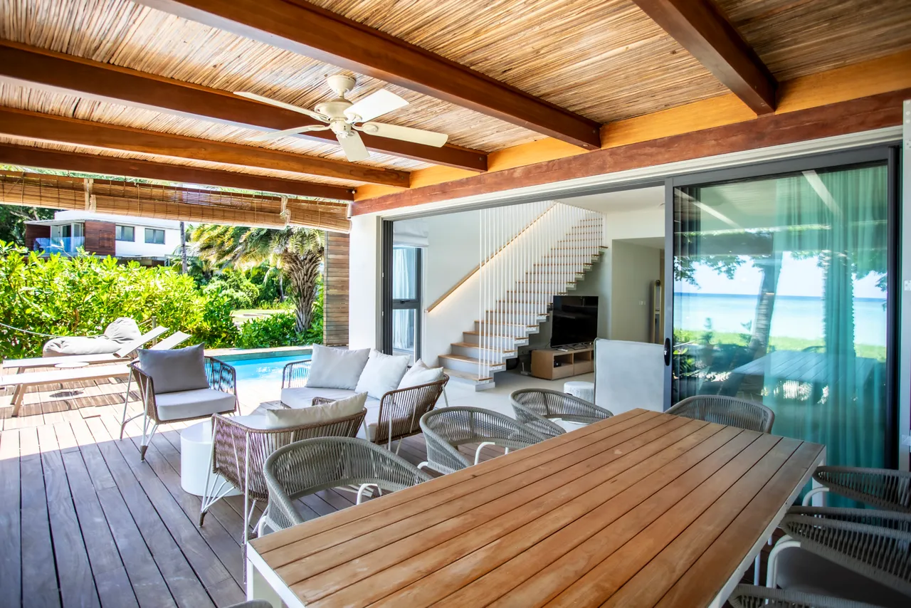 Teintes neutres du mobilier en rotin et bois sur la terrasse. Espace repas convivial avec vue sur la piscine. Grandes fenêtres laissant entrer la lumière naturelle. Accès direct à l'extérieur et à la plage.