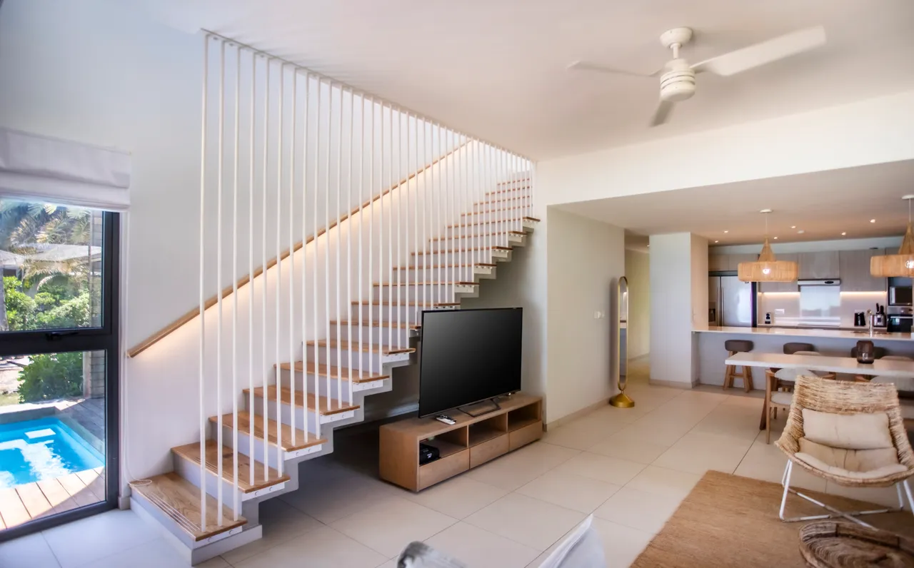 Escalier en bois clair menant aux niveaux supérieurs. Espace ouvert avec vue sur la piscine. Mobilier en rotin et table en bois ajoutent une touche contemporaine. Luminaires en osier apportent une ambiance chaleureuse.