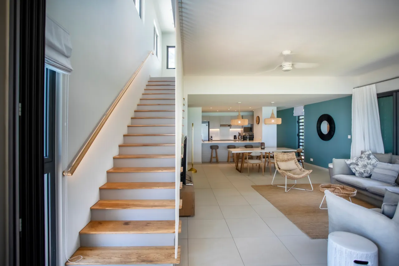 Lumière naturelle abondante dans le salon avec accès direct à l'extérieur. Mobilier contemporain et agencement ouvert créent un espace convivial. Escalier en bois clair menant aux niveaux supérieurs. Décoration sobre et fonctionnelle avec des touches de couleur.