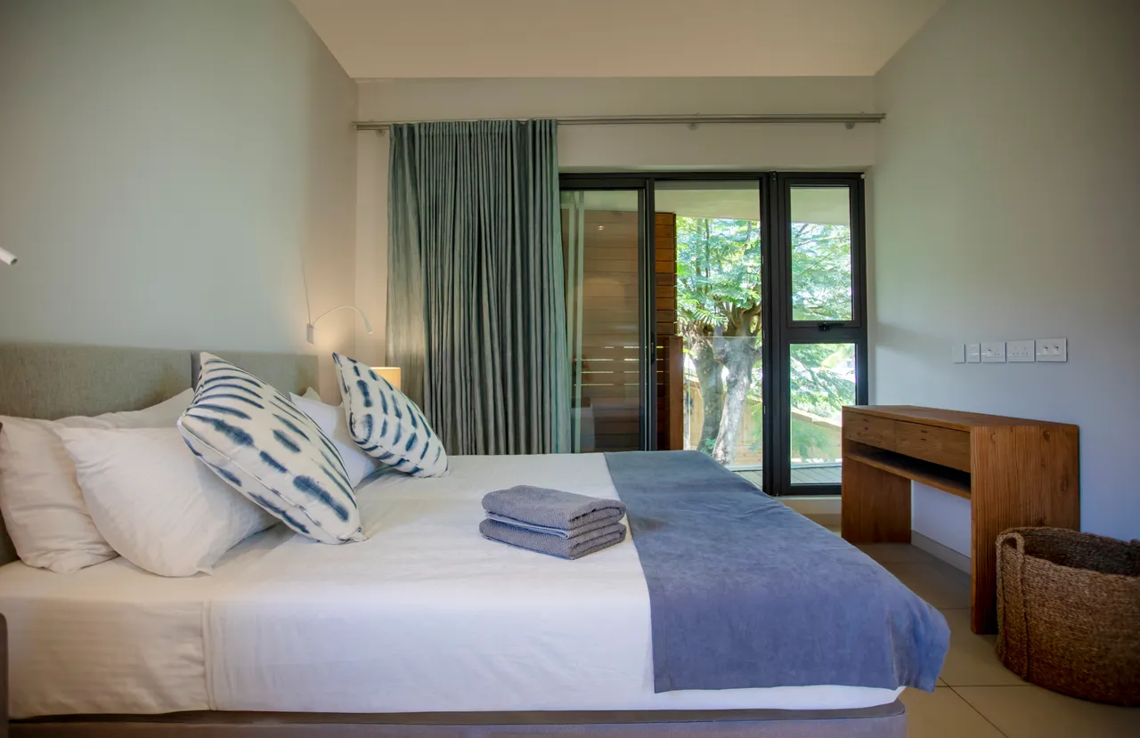 Chambre lumineuse avec accès direct à une terrasse privée. Mobilier en bois et textile clair. Grandes fenêtres offrant une vue sur l'extérieur. Espace de rangement fonctionnel.