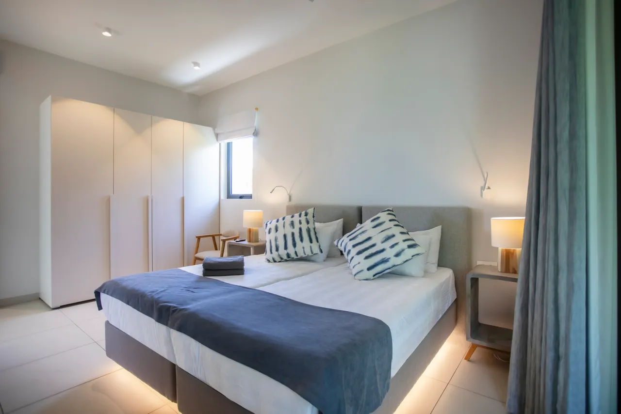 Chambre spacieuse avec lit double et espace de rangement intégré. Grandes fenêtres offrant une vue sur l'extérieur. Éclairage doux avec lampes de chevet. Mobilier en bois ajoutant une touche naturelle.