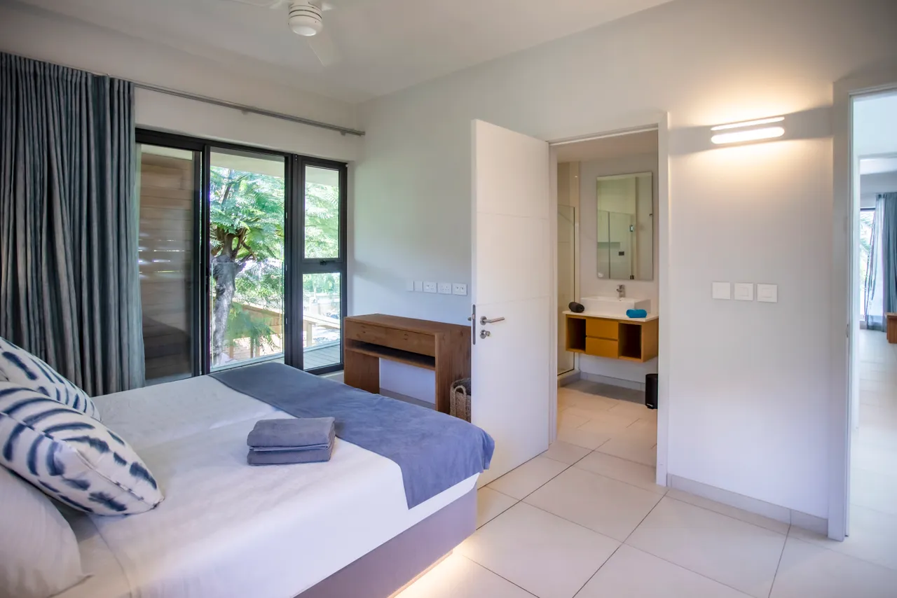 Teintes claires dominent la chambre, créant une atmosphère apaisante. Accès direct à la terrasse privée par des baies vitrées. Mobilier en bois et textile léger complètent l'aménagement. Salle de bain attenante avec éclairage naturel.