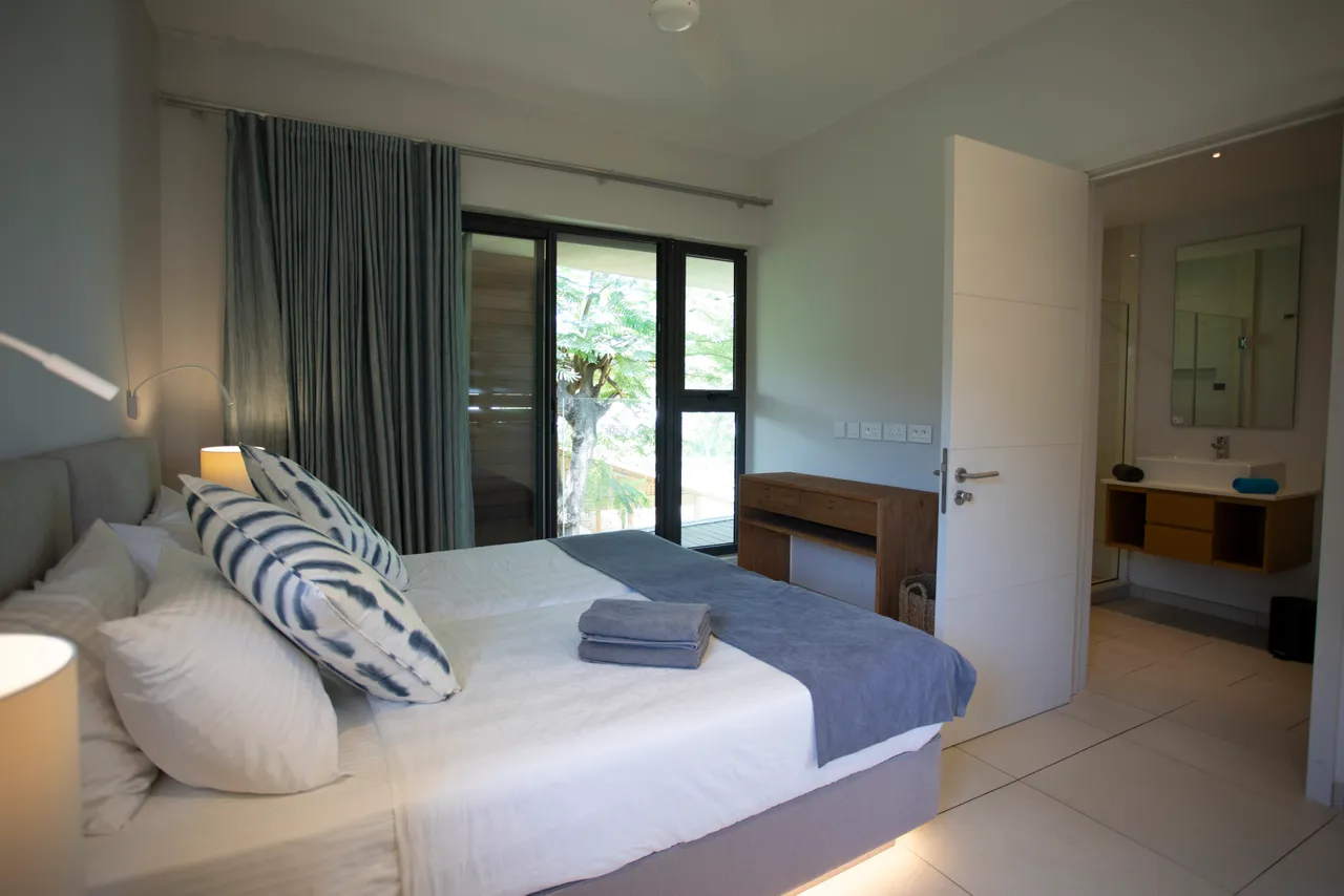 Sol carrelé et mobilier en bois clair dans la chambre. Textiles doux ajoutent une touche de confort. Fenêtres offrant une vue sur l'extérieur et accès à la lumière naturelle. Bureau fonctionnel intégré à l'aménagement. Salle de bain attenante accessible depuis la chambre.