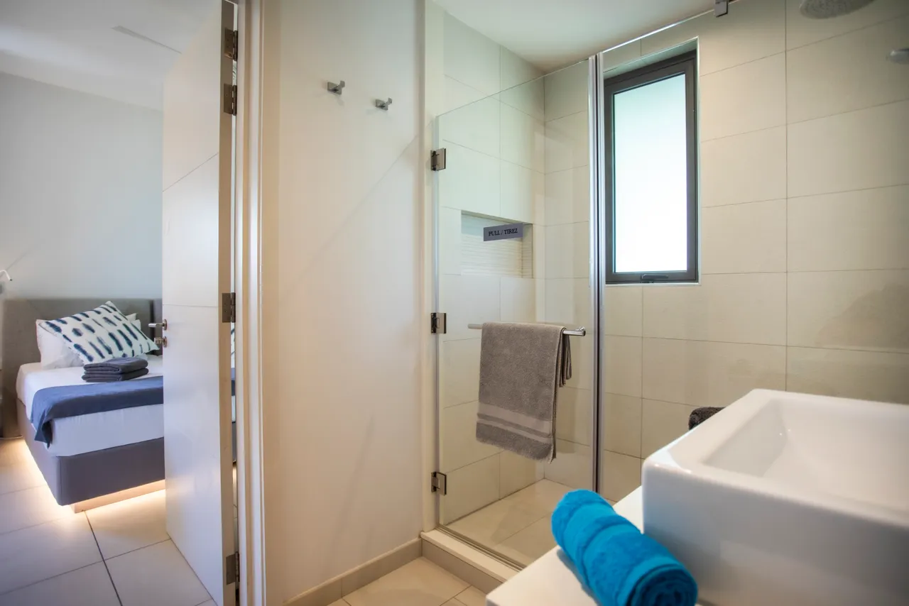 Teintes neutres et carrelage clair dans une salle de bain moderne. Douche à l'italienne avec accès à la lumière naturelle. Rangement intégré et décoration épurée pour une utilisation pratique. Éclairage doux agrémente l'espace fonctionnel.