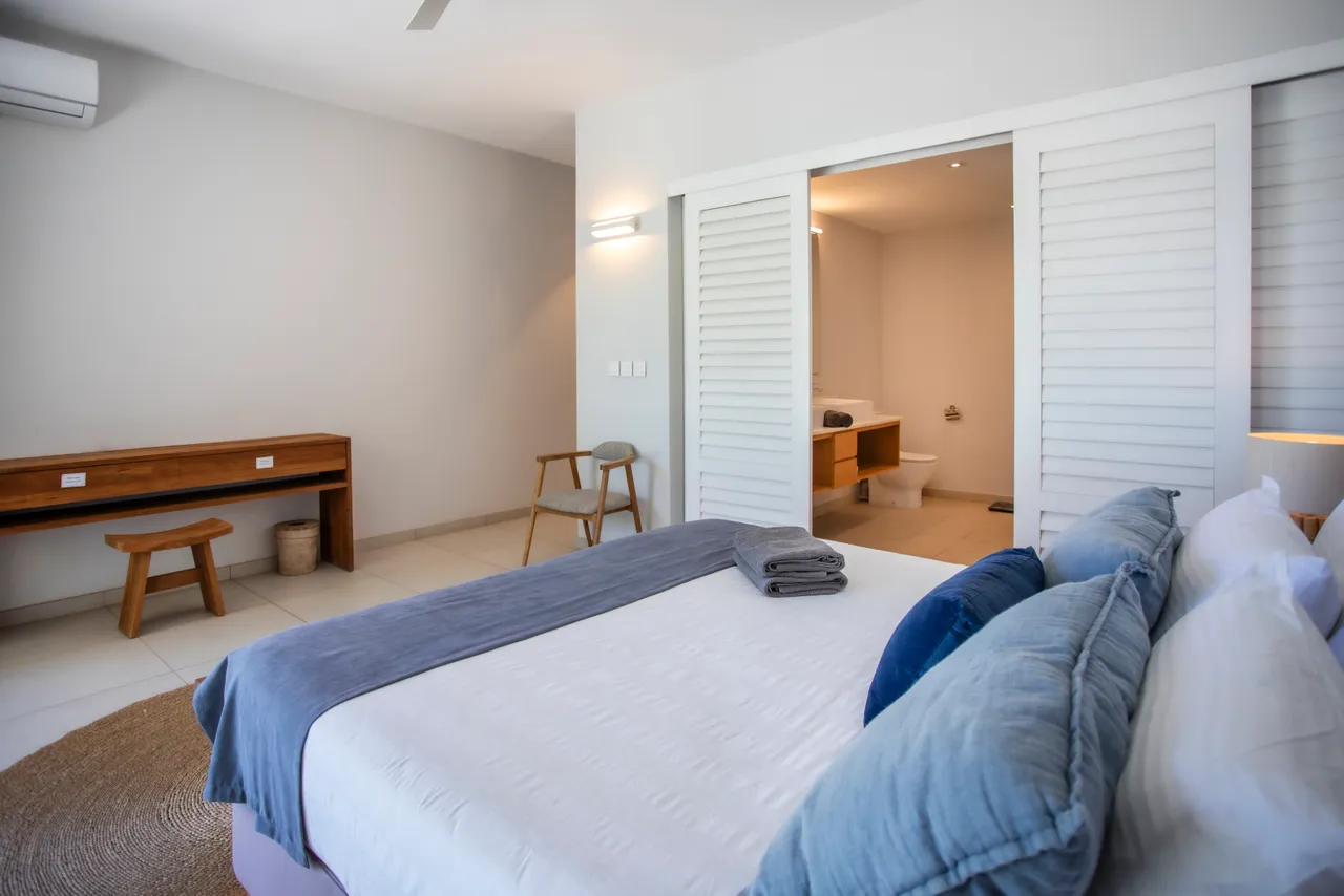 Chambre avec lit double et textile doux en lin. Bureau en bois clair et rangement discret. Fenêtres offrant une vue sur l'extérieur. Éclairage naturel grâce à un agencement ouvert. Accès direct à une salle de bain attenante.