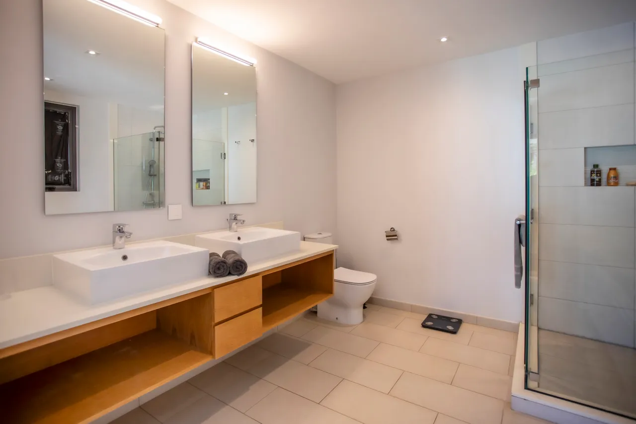 Salle de bain équipée de deux vasques et d'une douche à l'italienne. Éclairage naturel grâce à une fenêtre. Rangement pratique sous les lavabos en bois. Sol carrelé et décoration minimaliste.