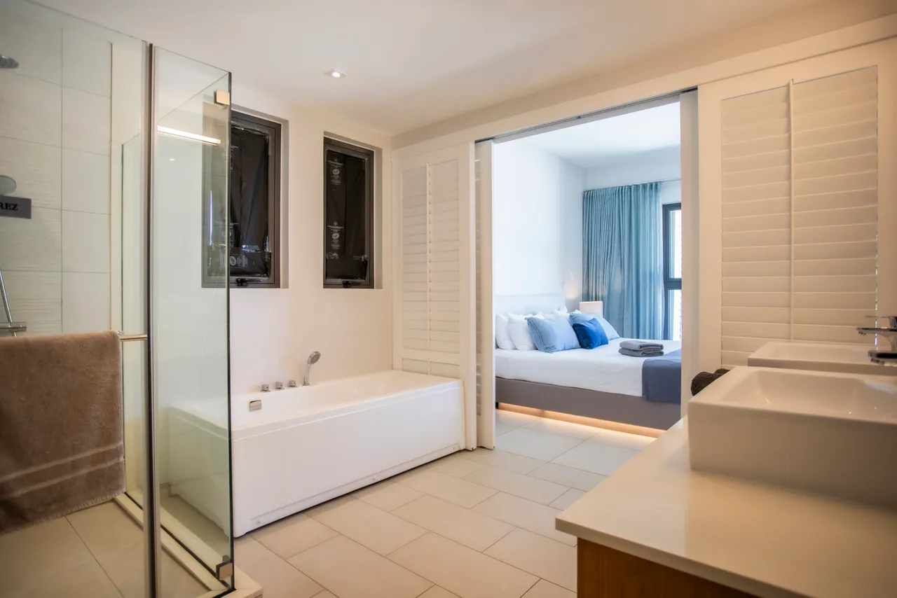 Salle de bain lumineuse avec baignoire et douche séparée. Rangement sous les vasques en bois. Sol en carrelage clair et décoration moderne. Éclairage naturel apporté par deux fenêtres.