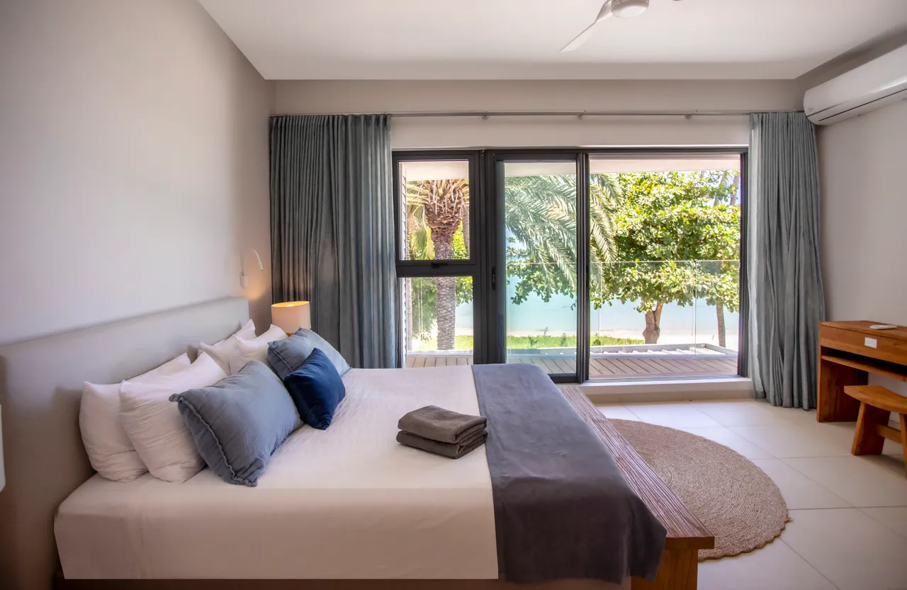 Chambre avec lit double et vue sur la mer à travers de grandes fenêtres. Mobilier en bois et textiles doux créent un espace apaisant. Accès direct à l'extérieur depuis la chambre. Éclairage naturel et aération assurés par un agencement ouvert.