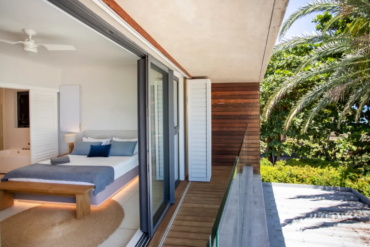 Chambre avec lit double et accès direct à un balcon. Grandes fenêtres offrant une vue sur la végétation. Mobilier en bois et textiles neutres pour une ambiance simple. Ventilation assurée par un ventilateur de plafond.