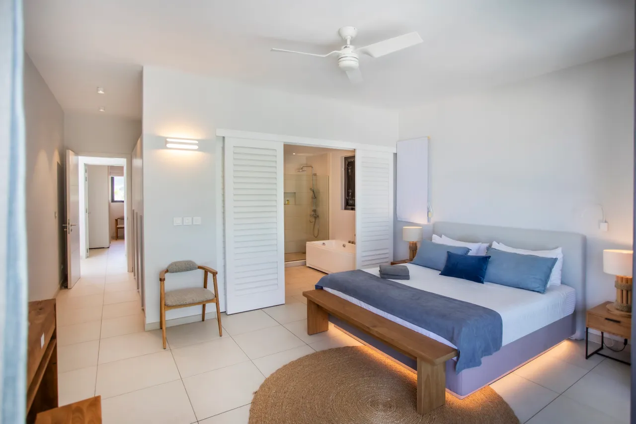 Chambre spacieuse avec lit double et accès à une salle de bain attenante. Mobilier en bois et textiles doux pour un aménagement minimaliste. Éclairage naturel assuré par de grandes fenêtres. Ventilateur de plafond pour un confort supplémentaire.