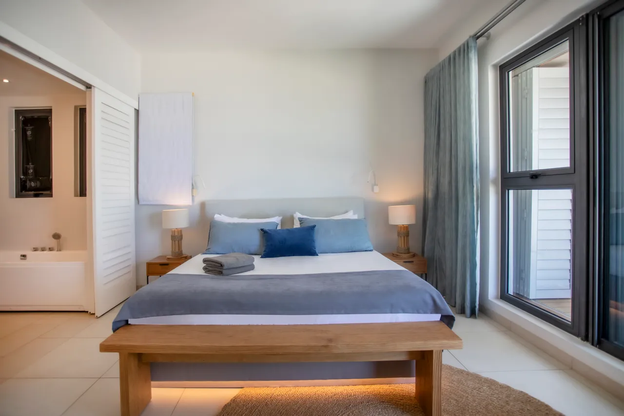 Chambre avec lit double et linge de lit en coton. Deux tables de nuit en bois avec lampes. Éclairage naturel grâce à de grandes fenêtres. Accès à une salle de bain attenante.
