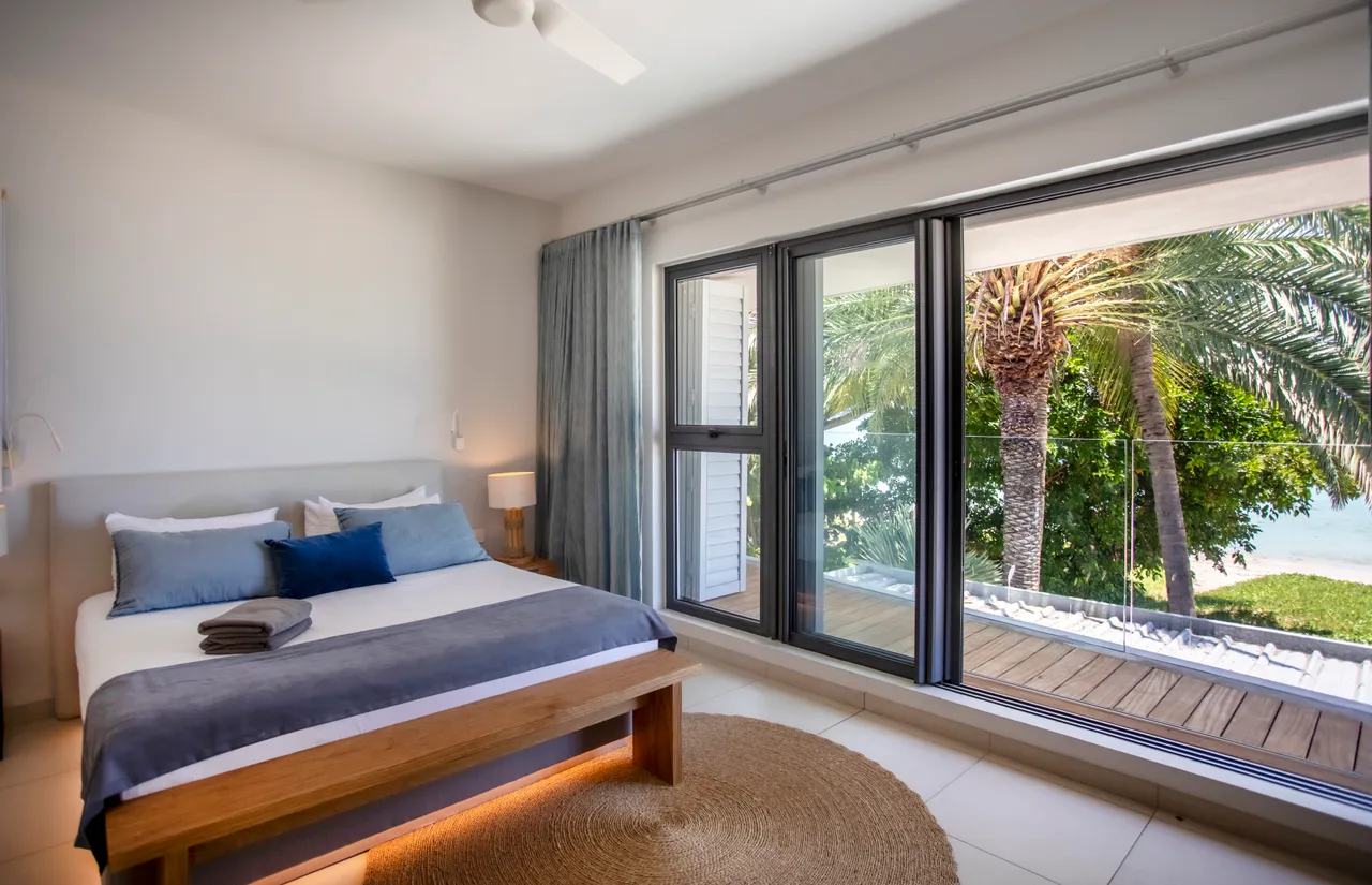 Chambre avec lit double et accès à une terrasse extérieure. Grandes fenêtres offrant une vue sur la végétation. Mobilier en bois et décor simple. Éclairage naturel et ventilation grâce à un ventilateur de plafond.