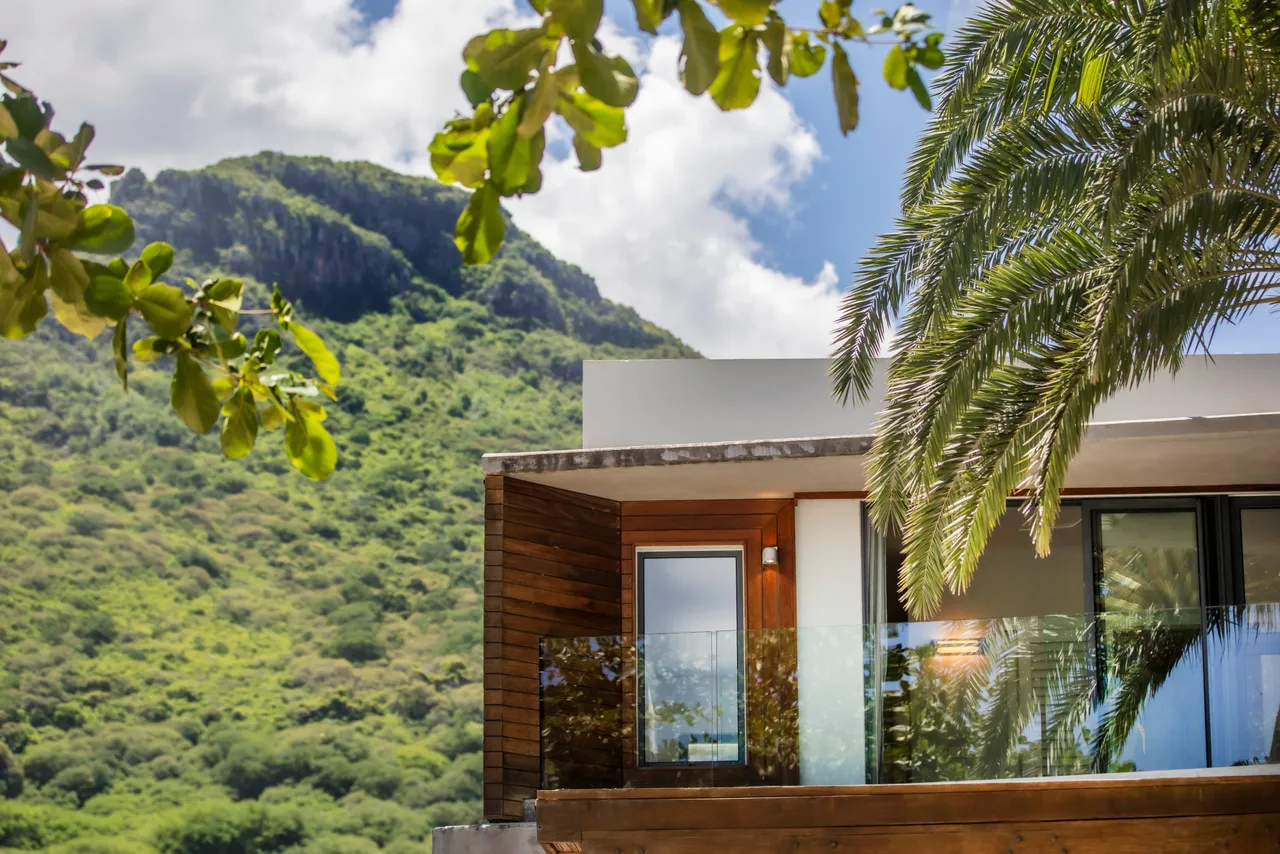 Villa avec terrasse en bois offrant une vue sur la montagne. Grandes fenêtres pour un éclairage naturel. Végétation tropicale apportant de l'ombre. Balustrade en verre pour une vue dégagée.