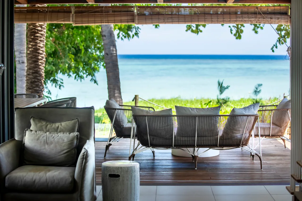 Terrasse en bois avec vue sur la mer et mobilier confortable. Espace extérieur idéal pour se détendre. Éclairage naturel grâce à des fenêtres ouvertes. Végétation tropicale apportant de l'ombre.