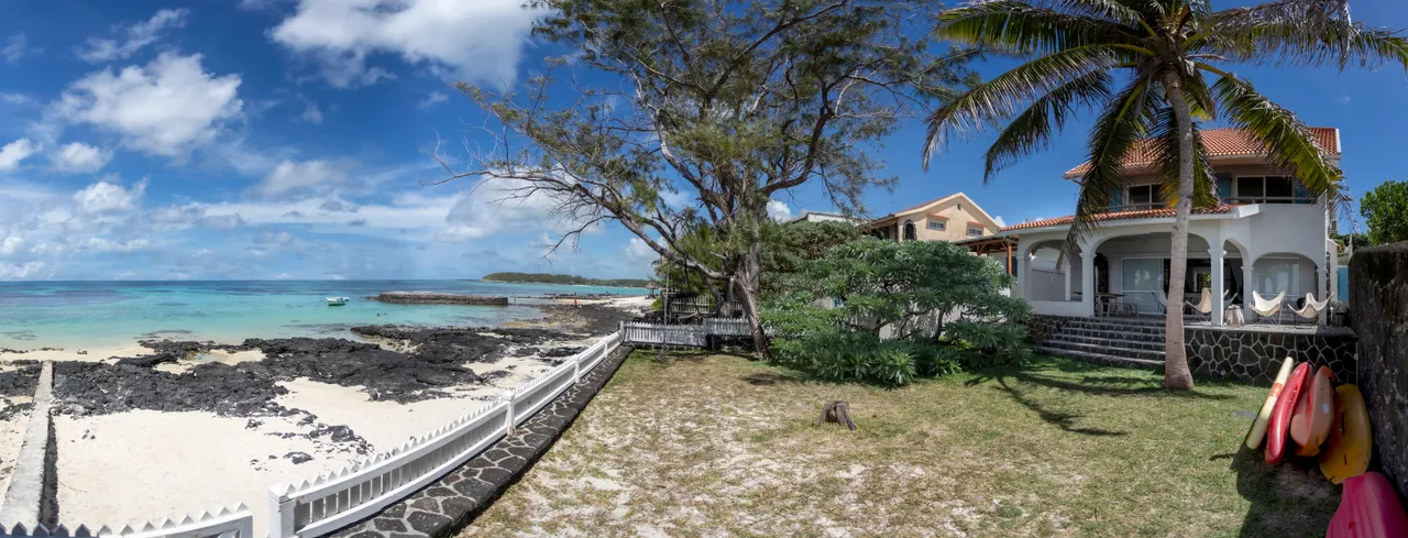Galerie. Accès direct à la plage depuis le jardin avec rochers visibles. Végétation tropicale bien entretenue.