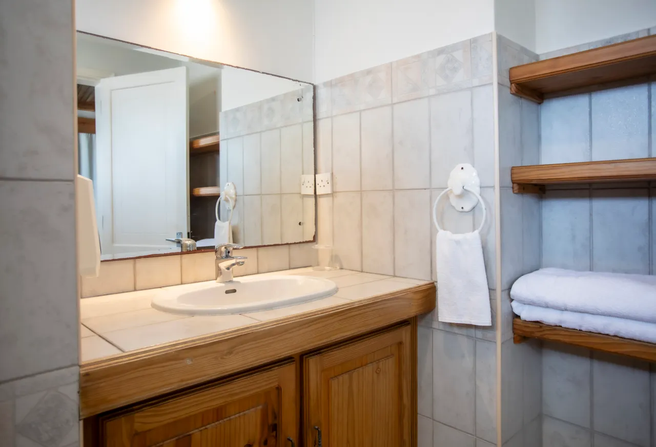 Murs en faïence claire et meuble en bois pour un rangement pratique. Lavabo encastré avec miroir et éclairage fonctionnel. Serviettes blanches bien disposées sur l'étagère.
