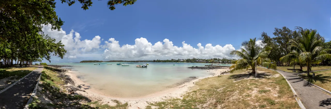 Plage de sable avec eaux claires et bateaux amarrés au loin. Palmiers bordent le rivage.