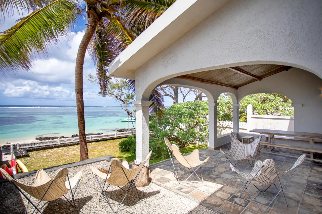 Espace extérieur couvert avec chaises en tissu et vue sur la mer. Sol en pierres naturelles et palmier à proximité.
