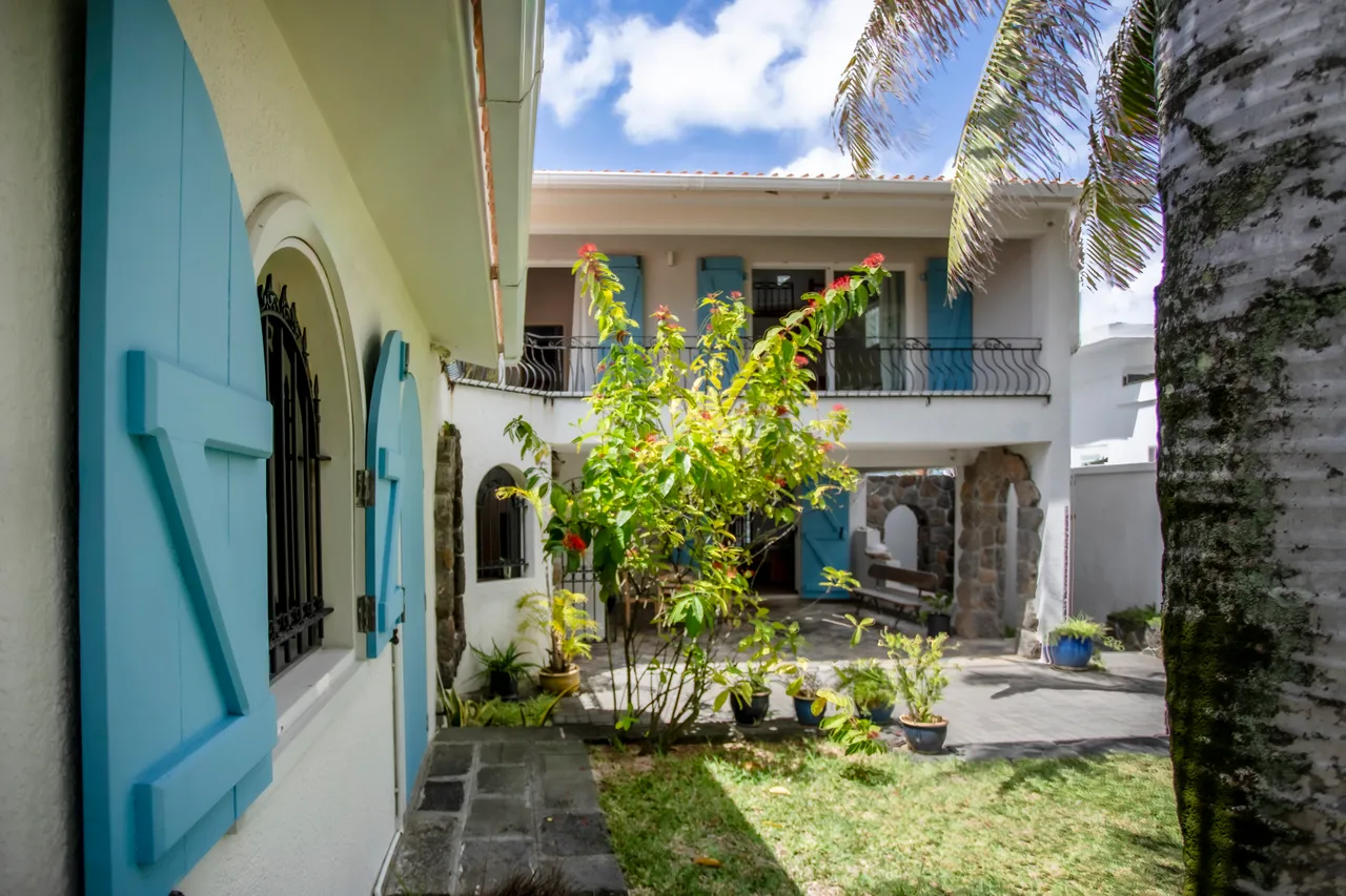 Espace extérieur avec jardin et plantes tropicales, offrant une atmosphère relaxante. Maison à deux niveaux avec volets bleus et accès direct au jardin.