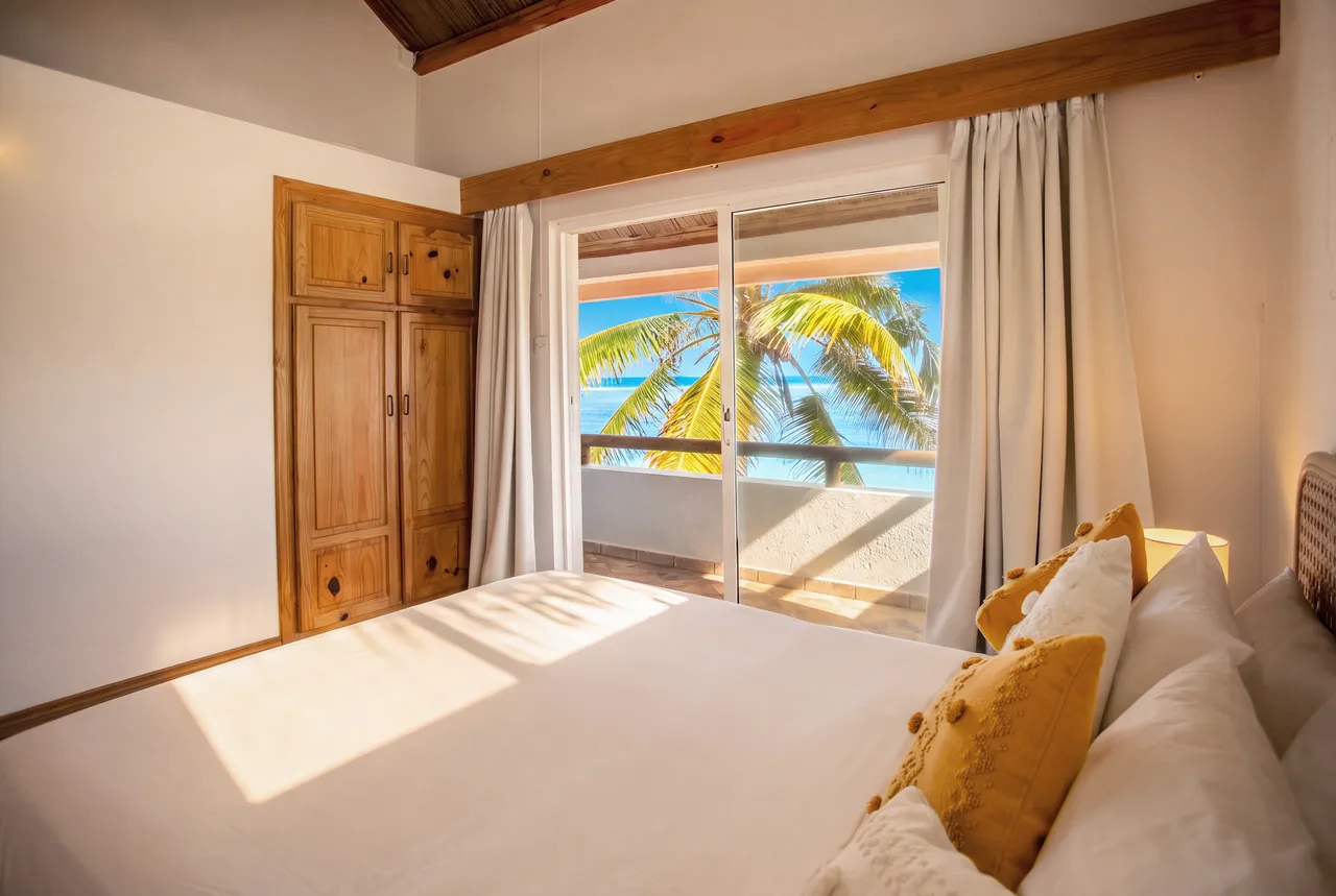 La lumière traverse la baie vitrée, avec des palmiers visibles depuis le lit.