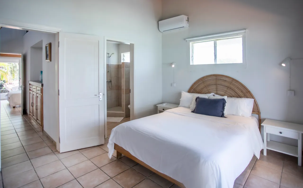 Chambre. Lit double avec tête de lit en rotin et rangements pratiques. Lumière naturelle grâce aux fenêtres.